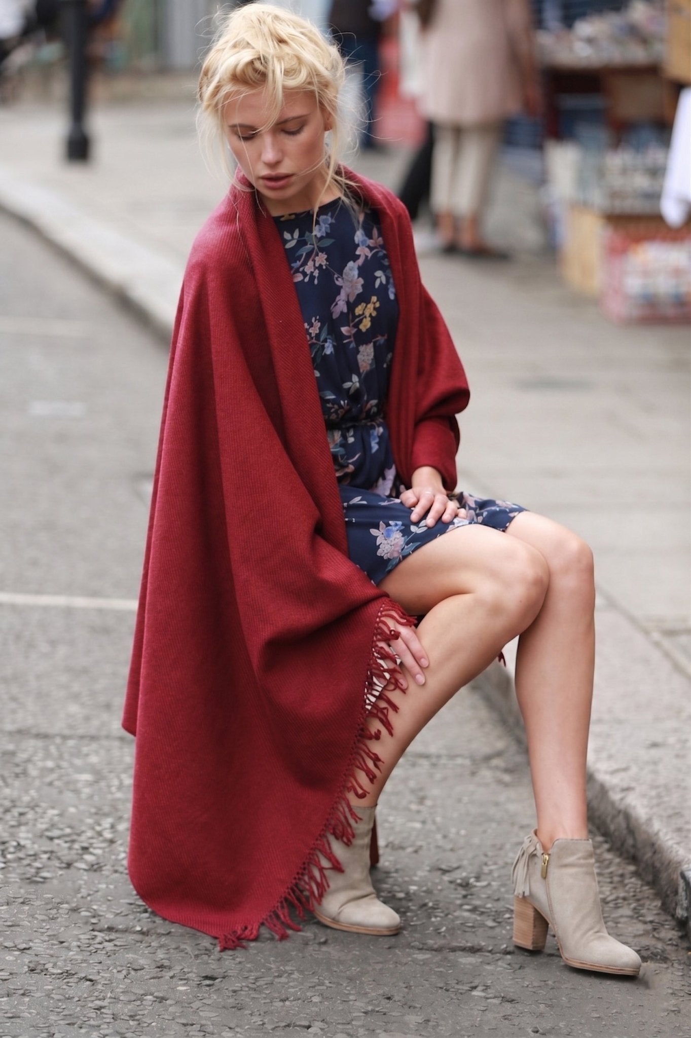 Woman sitting on a street wearing a red wool shawl and floral dress.